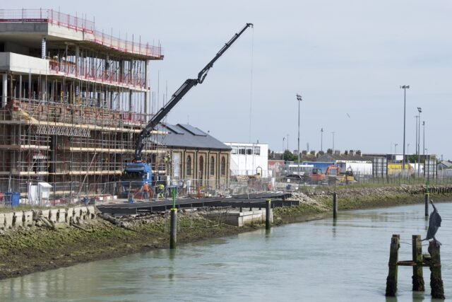 A view across the River Ouse in Newhaven, towards a construction site on the opposite bank. A mobile crane lowers a steel girder into place on a structure that comes out over the river bank onto three piles at the edge of the water. Behind is an old brick building surrounded by scaffolding.