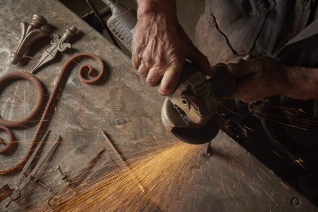 A top-down view of an old metal workbench. A person using an angle grinder, hands visible, sends a shower of sparks across the bench. Scrolled metalwork and finials sit on the bench alongside.