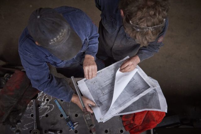 In a concrete-floored workshop, a top-down view of two men in denim overalls observing printouts of a construction plan.